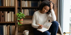 Woman sits on wooden stairs by a window, writing in a notebook with a pencil. She looks thoughtful, resting her head on her hand—perhaps contemplating the missing step in manifesting her dreams. Bookshelves, a plant, and a mug are nearby.