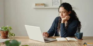 A person sits at a desk using a laptop, pondering why backwards manifesting exists, with an open notebook, a mug, and potted plants nearby.