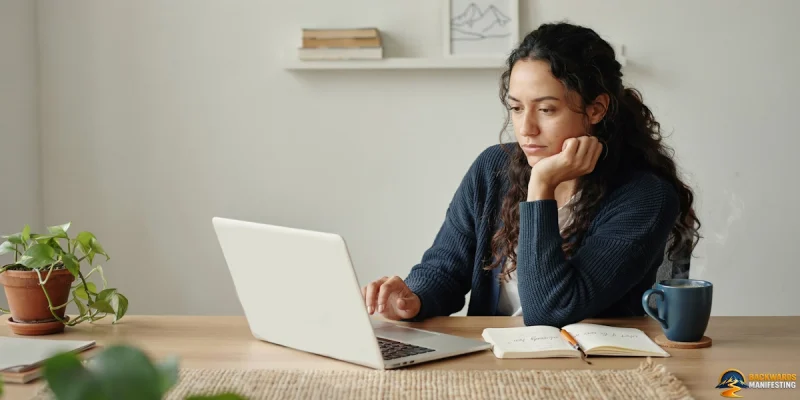 A person sits at a desk using a laptop, pondering why backwards manifesting exists, with an open notebook, a mug, and potted plants nearby.